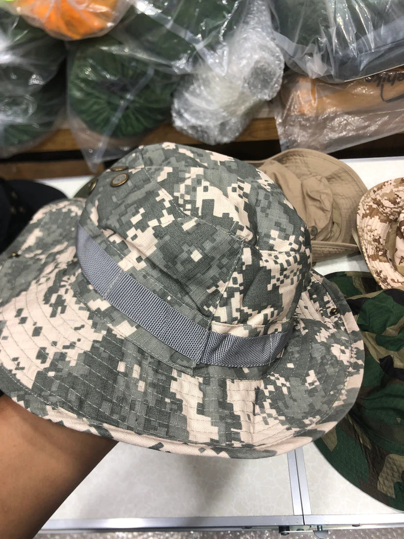 Camouflage-patterned hat with a gray strap held by a hand, with more hats in the background.