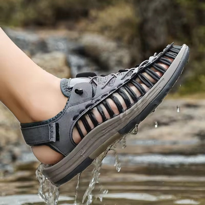Gray sandal with mesh design worn by a person stepping into water, with a natural background.