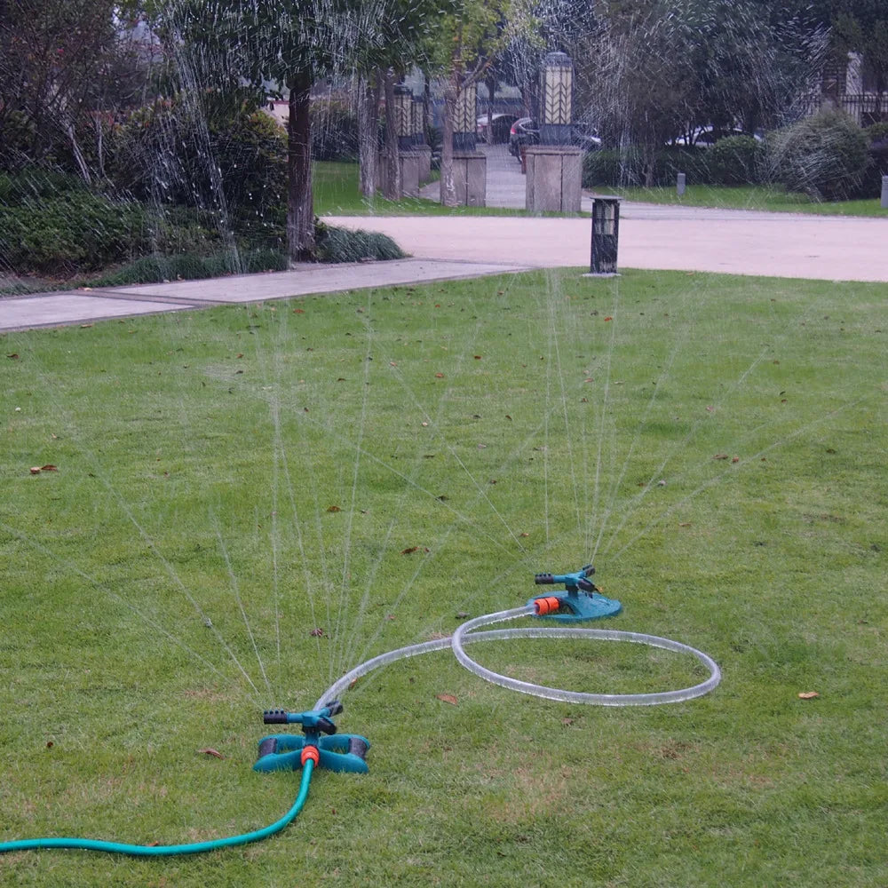 Sprinkler system watering a grassy area with a driveway and trees in the background.