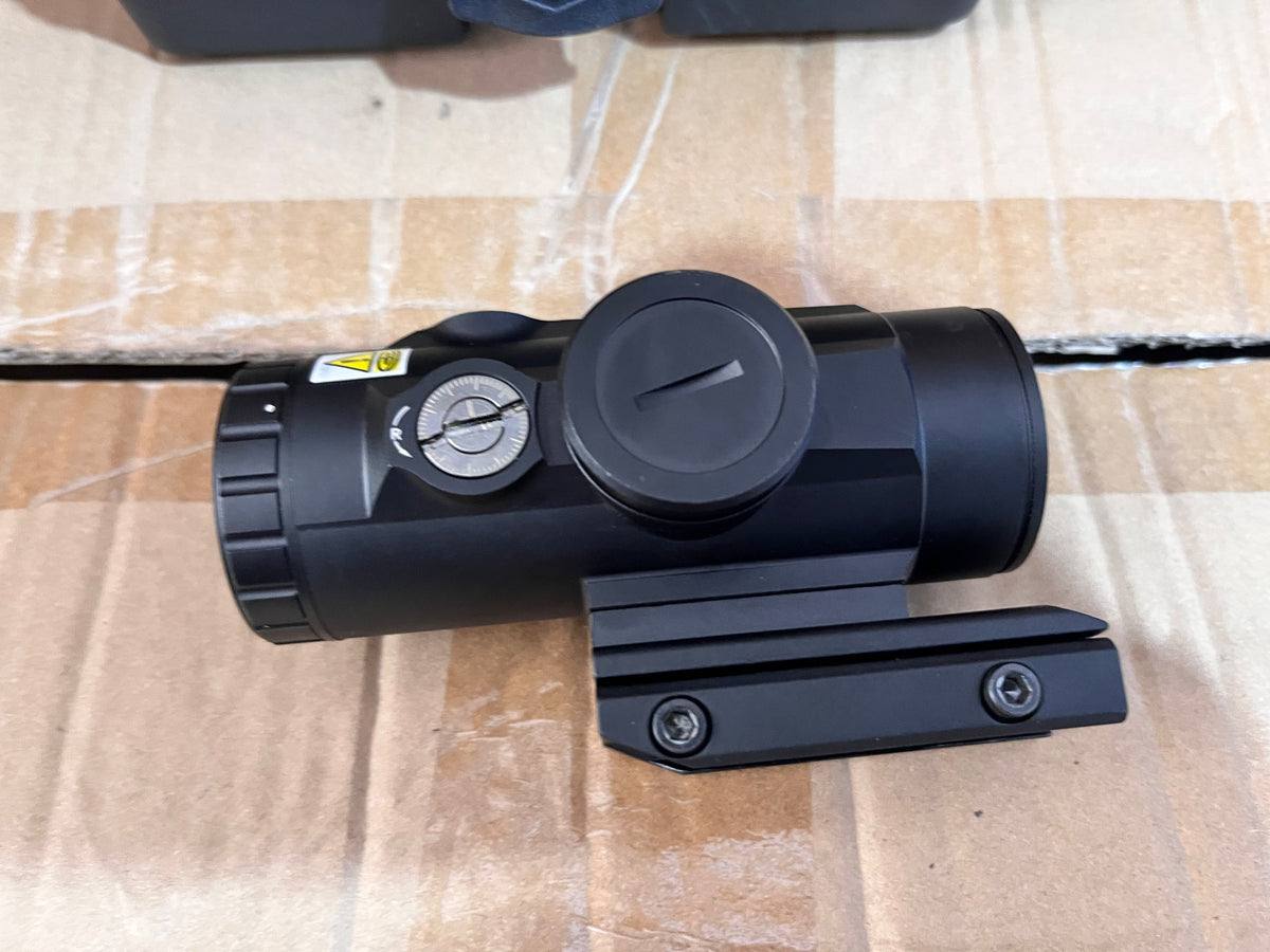 A black tactical rifle scope with a mounting plate, photographed on a brown cardboard box.